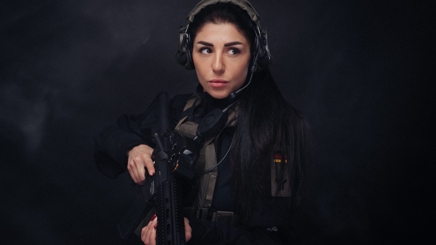 Schützin mit Gehörschutz und Plattenträger, die Langwaffe im Anschlag, vor dunklem Grund