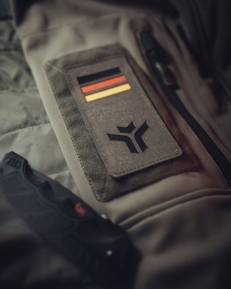 Rechteckiger Patch mit Deutschlandflagge und der Tacticool-Bildmarke, auf einer olivfarbenen Jacke